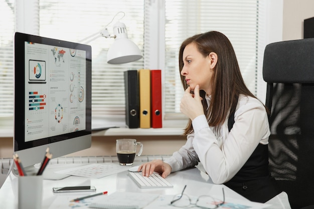 Bella sorridente donna d'affari dai capelli castani in giacca e occhiali seduto alla scrivania con una tazza di caffè e telefono cellulare, lavorando al computer moderno con documenti in ufficio leggero, mani sulla tastiera.