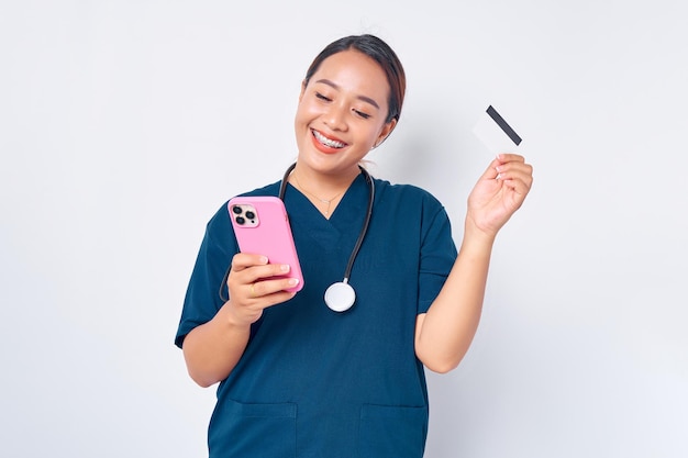 Bella sorridente giovane donna asiatica infermiera professionale che lavora indossando l'uniforme blu utilizzando il telefono cellulare e tenendo la carta di credito isolata su sfondo bianco Concetto di medicina sanitaria