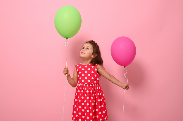 Bella splendida ragazza di compleanno graziosa di 4 anni in abito a pois rosa, tiene due palloncini multicolori, gioisce guardando un palloncino verde in mano, isolato su sfondo rosa con spazio di copia.