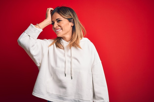 Bella sportiva di mezza età che indossa una felpa casual su sfondo rosso isolato sorridente fiducioso toccando i capelli con gesto della mano in alto posa attraente e alla moda