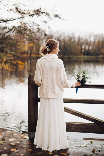 Bella sposa che sta da solo alla pera del fiume in vestito e maglione