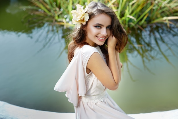 Bella sposa con capelli ricci lunghi in vestito da sposa che si siede vicino alla fontana al parco, foto del matrimonio, ritratto, primi piani.