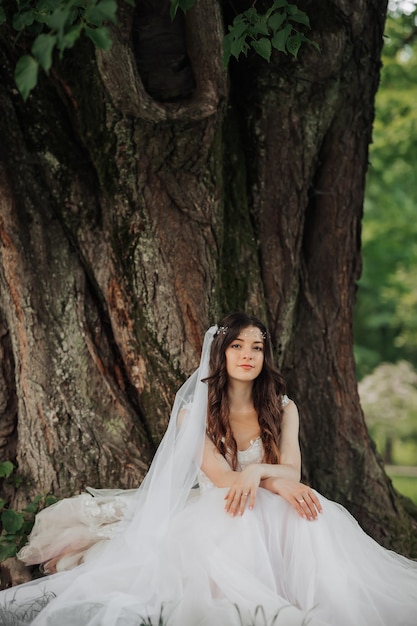 Bella sposa con i capelli lunghi e ricci in un vestito chic sorridendo guardando nell'obiettivo sotto un grande albero Ritratto della sposa Nozze primaverili Makeup naturale