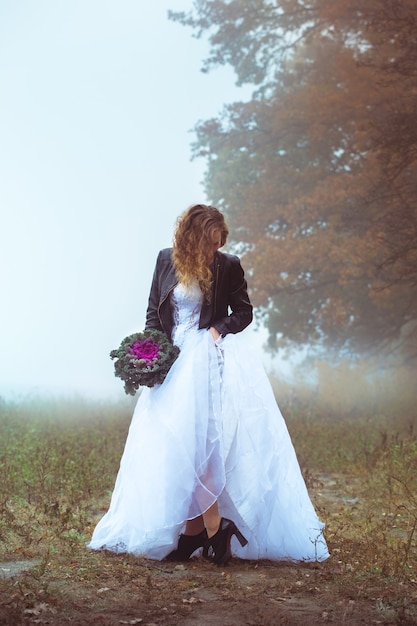 Bella sposa e una giacca di pelle con un bouquet sullo sfondo di un campo nebbioso