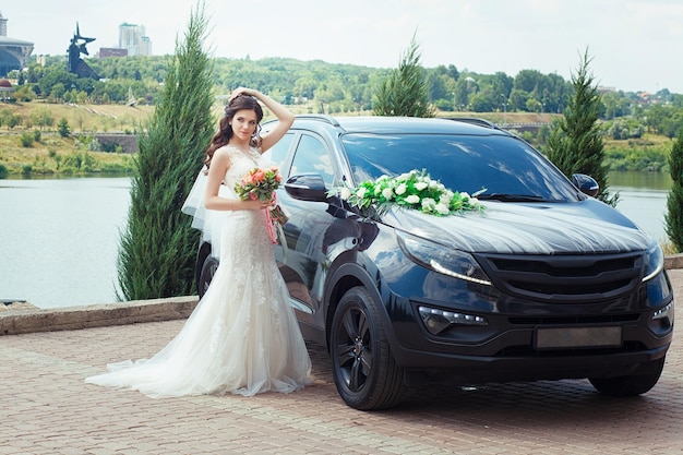 Bella sposa in abito da sposa si trova vicino all'auto in un parco cittadino. Bellissimo matrimonio.