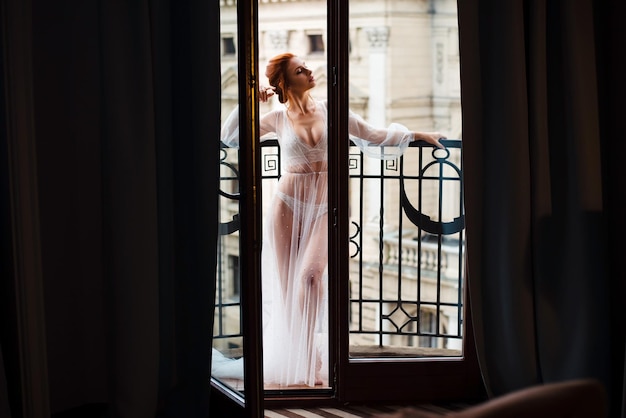 Bella sposa in piedi sul balcone in biancheria intima di pizzo bianco durante la sessione fotografica di matrimonio in un hotel moderno Donna sexy con trucco e acconciatura professionali