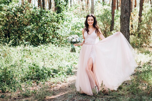 Bella sposa in un tenero abito rosa all'aperto, la ragazza tiene un bouquet, una ragazza in un abito lungo cammina in una pineta