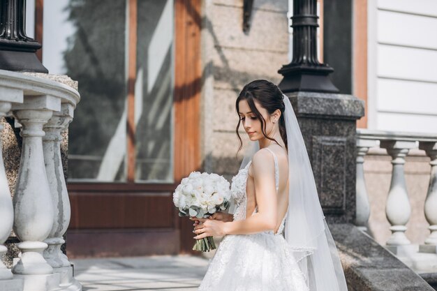 bella sposa sta in un vestito meraviglioso con un bouquet in mano vicino a una grande casa grigia