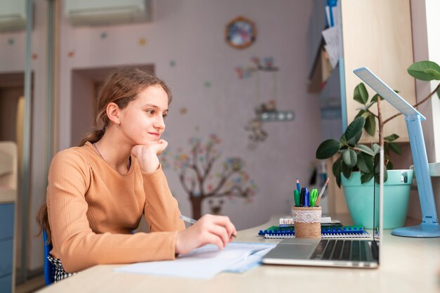 Bella studentessa che studia a casa facendo i compiti della scuola. Libri di formazione e taccuino sul tavolo. Formazione online a distanza