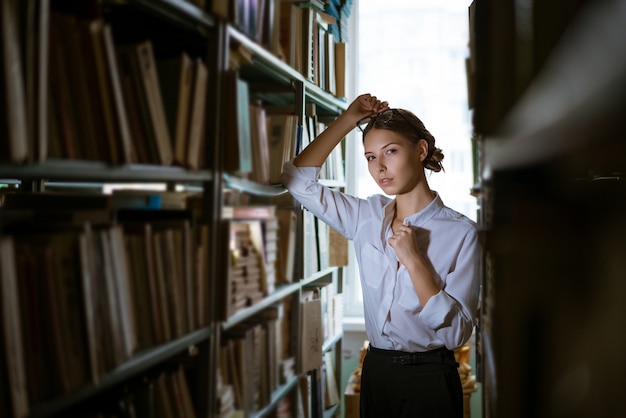 Bella studentessa con una camicia bianca si trova tra le file della biblioteca, scaffali per libri vale la pena di libri.