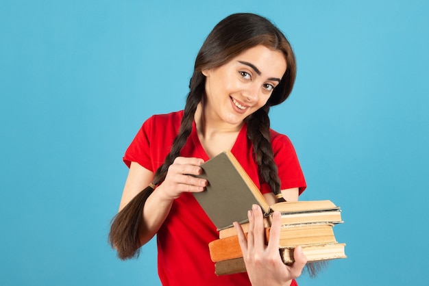 Bella studentessa in maglietta rossa leggendo attentamente il libro sulla parete blu.