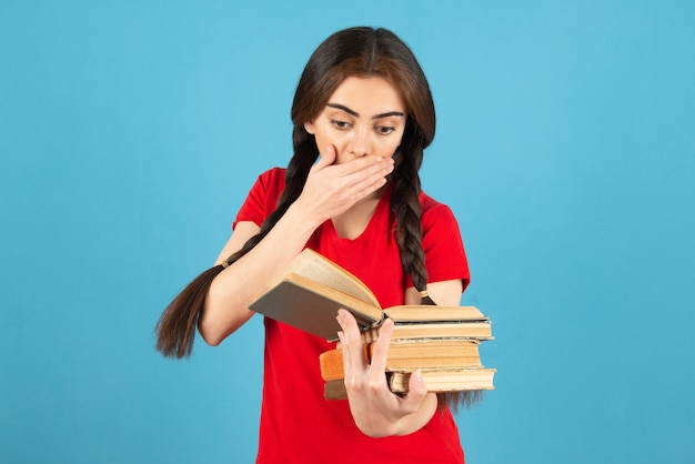 Bella studentessa nel libro di lettura della maglietta rossa con l'espressione scioccata.