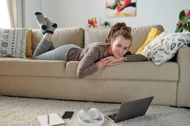 Bella studentessa sdraiata sul divano e ti guarda mentre prepara i compiti davanti al laptop a casa