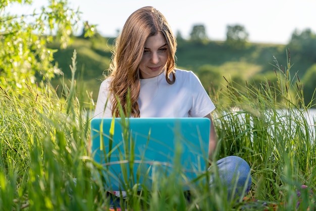 Bella studentessa si siede sull'erba in riva al lago con i laptop in grembo ed esamina con entusiasmo qualcosa