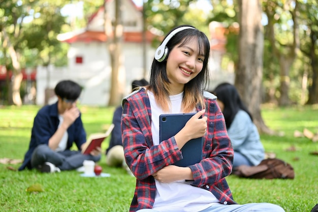 Bella studentessa universitaria asiatica che indossa le cuffie tenendo il suo tablet nel parco del campus