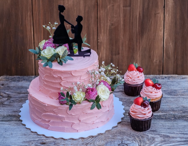 Bella torta nuziale a due livelli con crema di formaggio rosa, decorata con rose vive e una figura di coppia innamorata, su un tavolo di legno con cupcakes