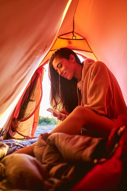 Bella viaggiatrice che invia messaggi di testo su smartphone e sorride mentre è seduta in una tenda turistica