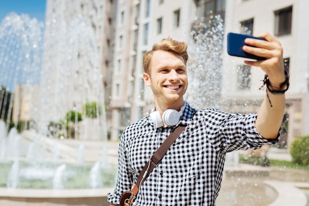 Bella vista. Felice l'uomo felice in piedi vicino alla fontana mentre sorride e prende selfie