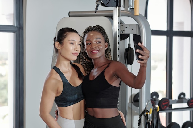 Belle amiche sorridenti che prendono selfie dopo essersi esercitati insieme in palestra