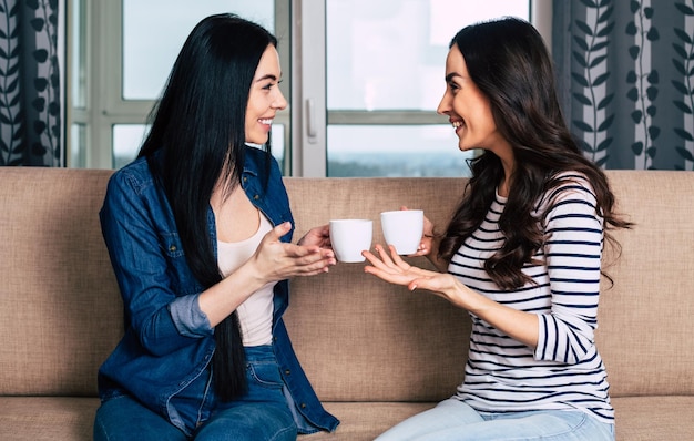 Belle amiche sorridenti in abiti casual seduti sul divano di casa a parlare e bere caffè o tè