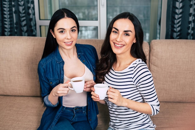 Belle amiche sorridenti in abiti casual seduti sul divano di casa a parlare e bere caffè o tè