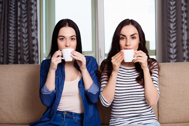 Belle amiche sorridenti in abiti casual seduti sul divano di casa a parlare e bere caffè o tè