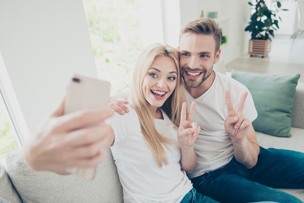 Belle coppie in magliette bianche casual e jeans a casa fanno selfie mostrando vsign