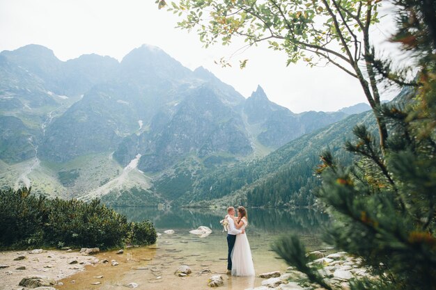 Belle coppie moderne vicino a un lago in montagna fanno foto di matrimonio