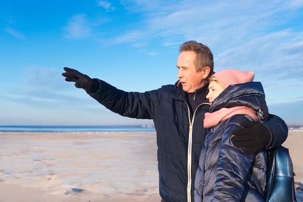 Belle coppie pensionate senior europee anziane nell'amore che cammina sulla spiaggia, mare in un giorno di inverno in vestiti caldi. L'uomo abbraccia la donna, la moglie, mostra, guarda, indica qualcosa in lontananza