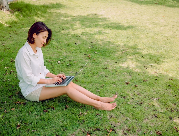 Belle donne asiatiche che indossano camicia bianca e shorts con il computer portatile che si siede sull'erba verde.