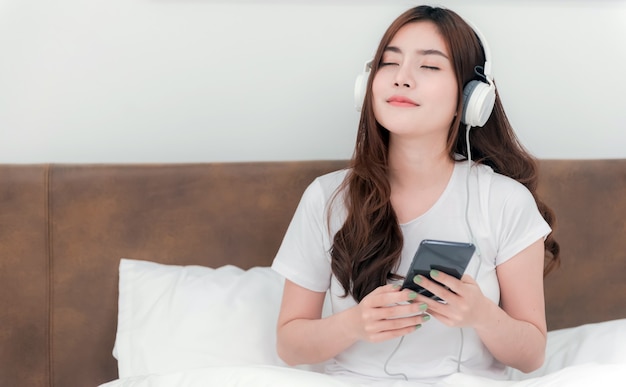 Belle donne asiatiche che indossano le cuffie che ascoltano la musica in streaming dai loro smartphone. Sul letto nella camera bianca