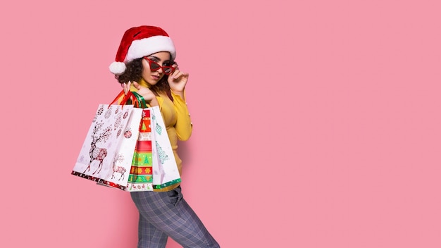 Belle donne che indossano un Natale luminoso che trasportano borse della spesa colorate su sfondo rosa Shopping natalizio e felice anno nuovo sconti sulle vendite nei negozi di tutto il mondo