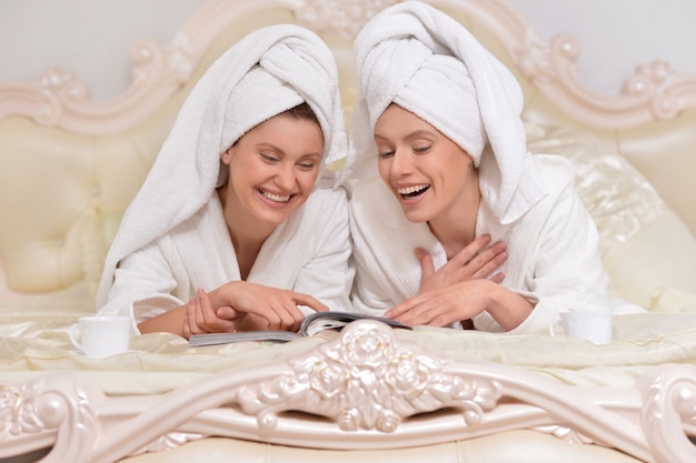 Belle giovani donne che indossano un accappatoio bianco leggendo una rivista a letto