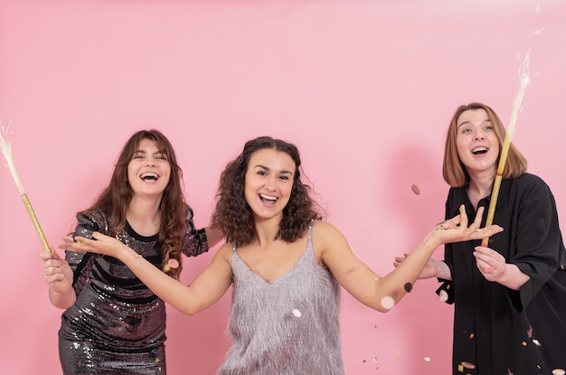 Belle ragazze allegre vestite a festa su sfondo rosa con fuochi d'artificio, candele e coriandoli, concetto di festa di capodanno.