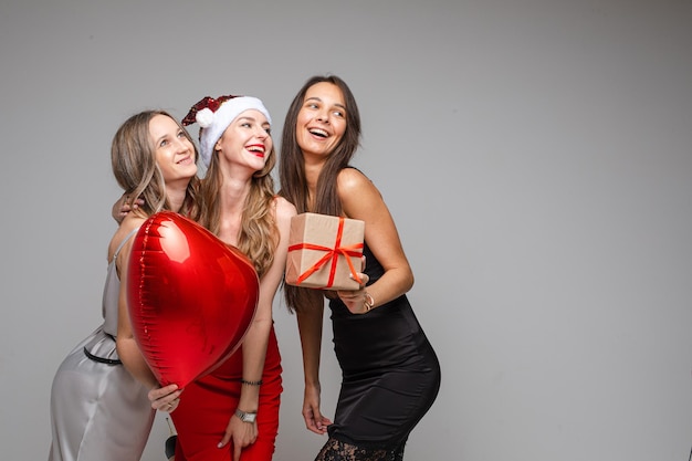 Belle ragazze amiche in abiti festivi che tengono regalo, palloncino rosso, champagne che celebrano le vacanze su sfondo grigio