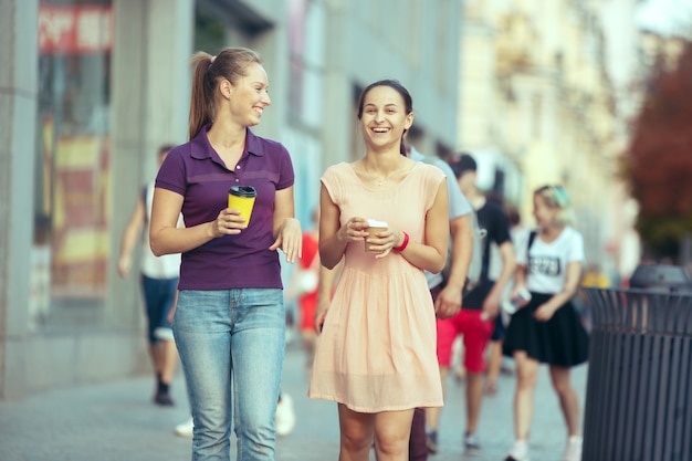 Belle ragazze che tengono una tazza di caffè di carta e si godono la passeggiata in città durante la giornata di sole estivo. Concetto di stile di vita