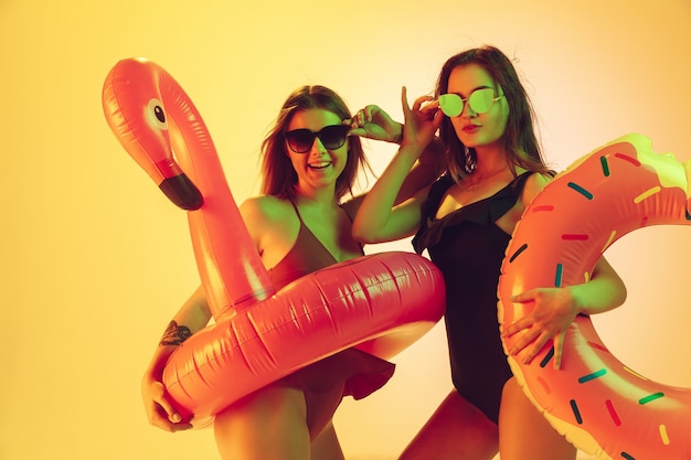 Belle ragazze in costumi da bagno alla moda isolati su sfondo giallo studio alla luce al neon. Estate, resort, moda e concetto di fine settimana. In posa in ciambella di gomma e fenicottero con emozioni luminose.