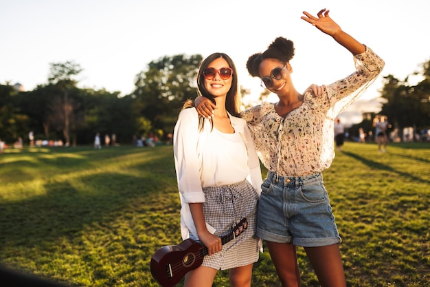 Belle ragazze sorridenti in occhiali da sole con piccola chitarra felicemente guardando a porte chiuse mentre trascorre del tempo insieme nel parco