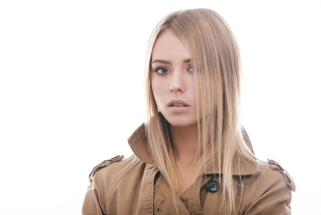 Bellezza alla moda. Ritratto di giovane donna attraente in cappotto che guarda l'obbiettivo mentre in piedi su sfondo bianco