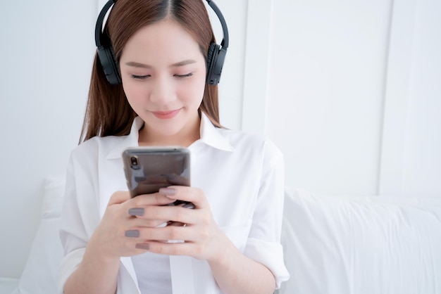 Bellezza asiatica femminile vestito bianco capelli lunghi indossare cuffia e utilizzare il tipo di ricerca smartphone sul letto bianco con vongole e allegro sfondo bianco camera da letto interna