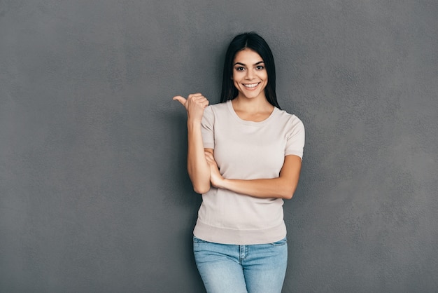 Bellezza che indica lo spazio della copia. Attraente giovane donna in abbigliamento casual che punta lontano e sorride mentre è in piedi su sfondo grigio