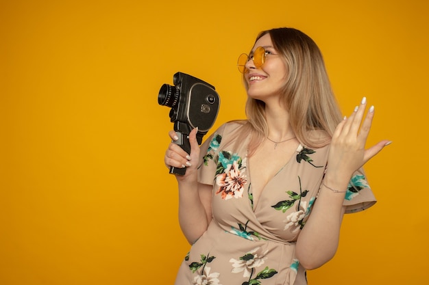 Bellezza con la cinepresa. Giovane donna allegra che tiene la cinepresa e posa su sfondo giallo