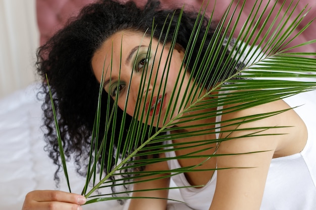 Bellezza, cura della pelle e concetto di persone - Close Up ritratto di bella donna di razza mista. ritratto di una donna africana, sdraiata su un letto bianco come la neve, guardando la telecamera. Ragazza e ramo di palma.