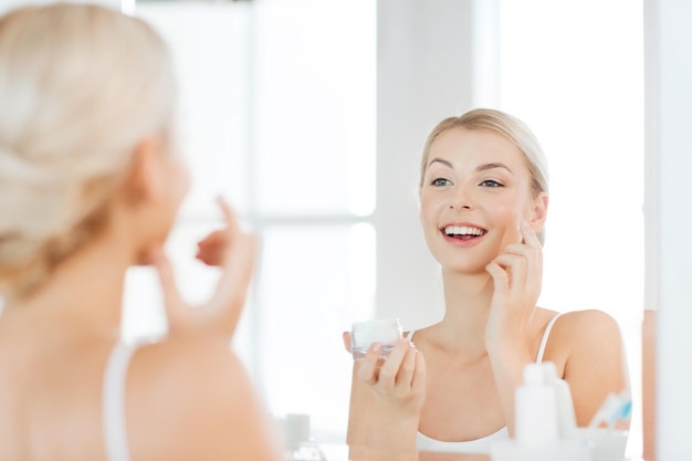 Bellezza, cura di pelle e concetto della gente - giovane donna sorridente che applica crema per affrontare e che guarda per rispecchiare a casa il bagno