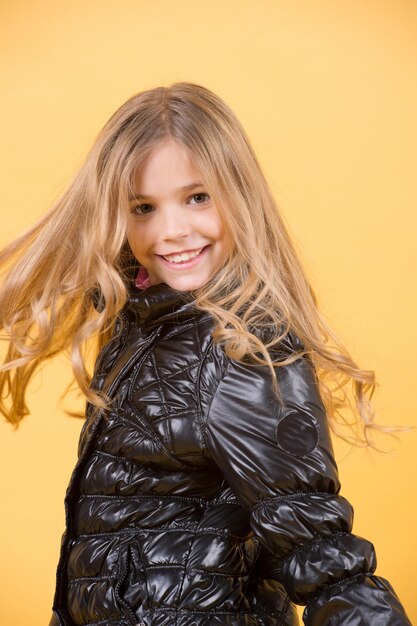 Bellezza del bambino, aspetto, acconciatura. Sorriso modello bambino con lunghi capelli biondi. Ragazza in cappotto nero su sfondo arancione. Moda, stile autunnale, tendenza. Concetto di infanzia felice.