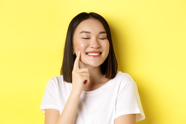 Bellezza e cura della pelle. Primo piano di giovane donna asiatica con corti capelli scuri, pelle sana e luminosa, sorridente e toccante fossette sulle guance, in piedi su sfondo giallo.