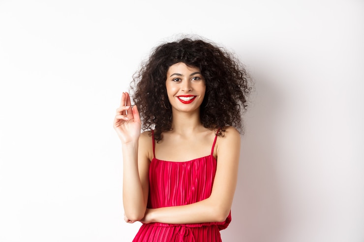 Foto bellezza e moda. donna sorridente con capelli ricci e trucco, indossa un abito rosso, agitando la mano in gesto di saluto, salutando, in piedi su sfondo bianco.
