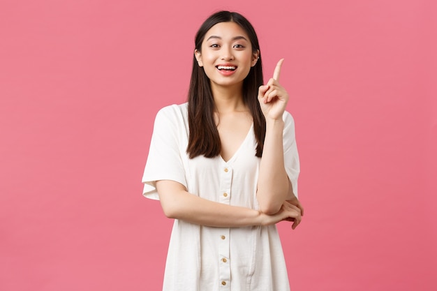 Bellezza, emozioni delle persone e concetto di svago estivo. Una bella ragazza asiatica eccitata in abito bianco ha un'idea, alza il dito indice e suggerisce una soluzione, racconta il suo piano, sfondo rosa