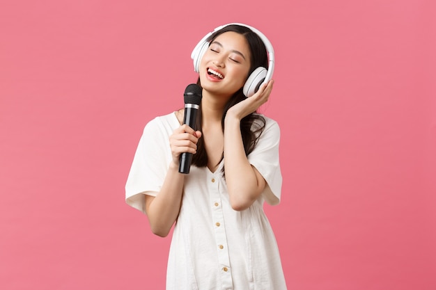 Bellezza, emozioni delle persone e concetto di tecnologia. Ragazza asiatica felice e spensierata che utilizza l'applicazione karaoke del telefono cellulare, canta nel microfono, ascolta musica in cuffia, sfondo rosa.