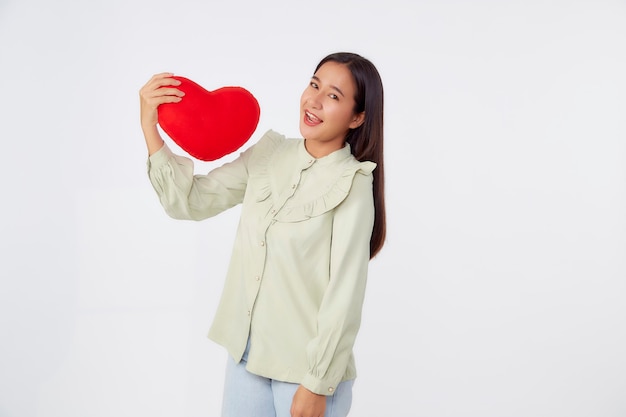 Bellezza giovane bruna asiatica donna che indossa la camicia verde in piedi tenere cuscino a forma di cuore rosso nella superficie dello studio isolato grigio chiaro